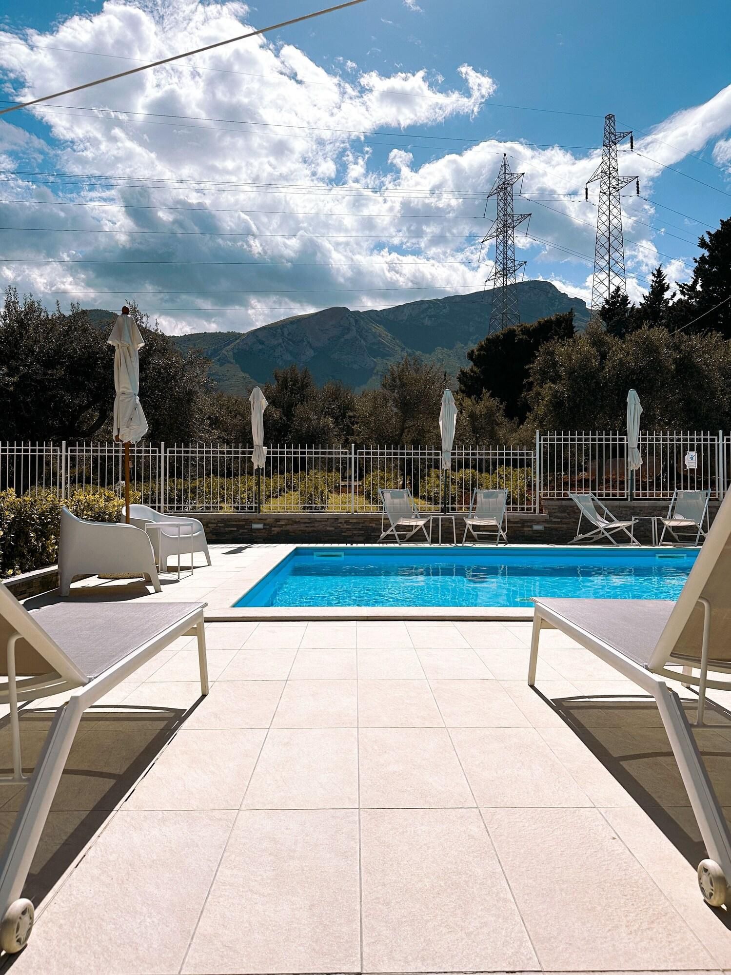Une piscine entourée d’une clôture, avec des chaises longues et des parasols, et en arrière-plan, des montagnes et des lignes électriques.