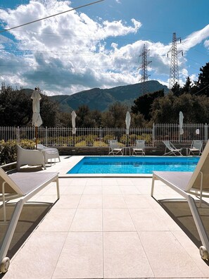 Une piscine entourée d’une clôture, avec des chaises longues et des parasols, et en arrière-plan, des montagnes et des lignes électriques.