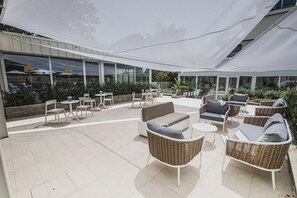 An outdoor patio area with wicker furniture, a white canopy, and a view of the building's interior through large windows.