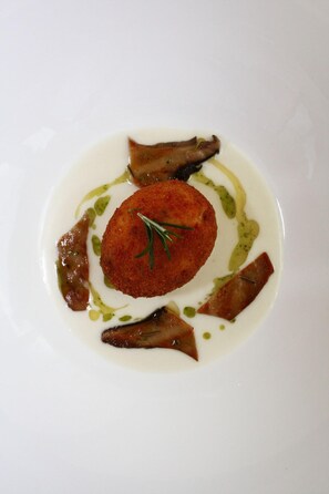 Une assiette blanche avec une seule portion ronde et panée au centre, entourée de tranches de champignons et nappée d’une sauce verte.