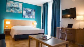 A hotel room with a bed, bedside lamps, a TV, a desk with a laptop, and two abstract paintings on the wall.