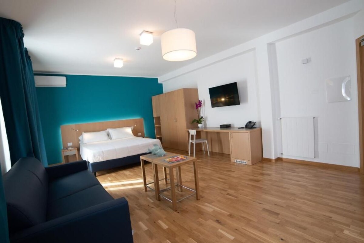 A modern hotel room with a bed, a desk, a chair, a small table, and a TV on the wall.