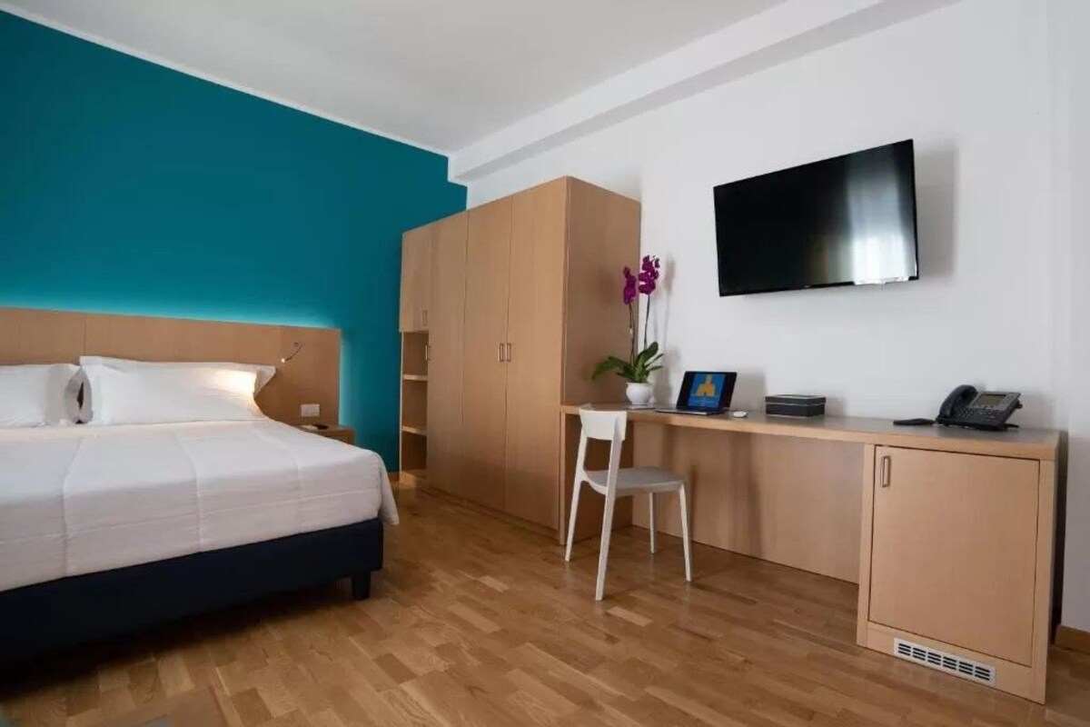 A hotel room with a bed, a desk, a chair, a TV, and a wardrobe.