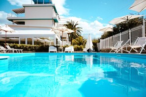 Une piscine entourée d’une clôture blanche et de verdure, avec des chaises longues blanches et des parasols.