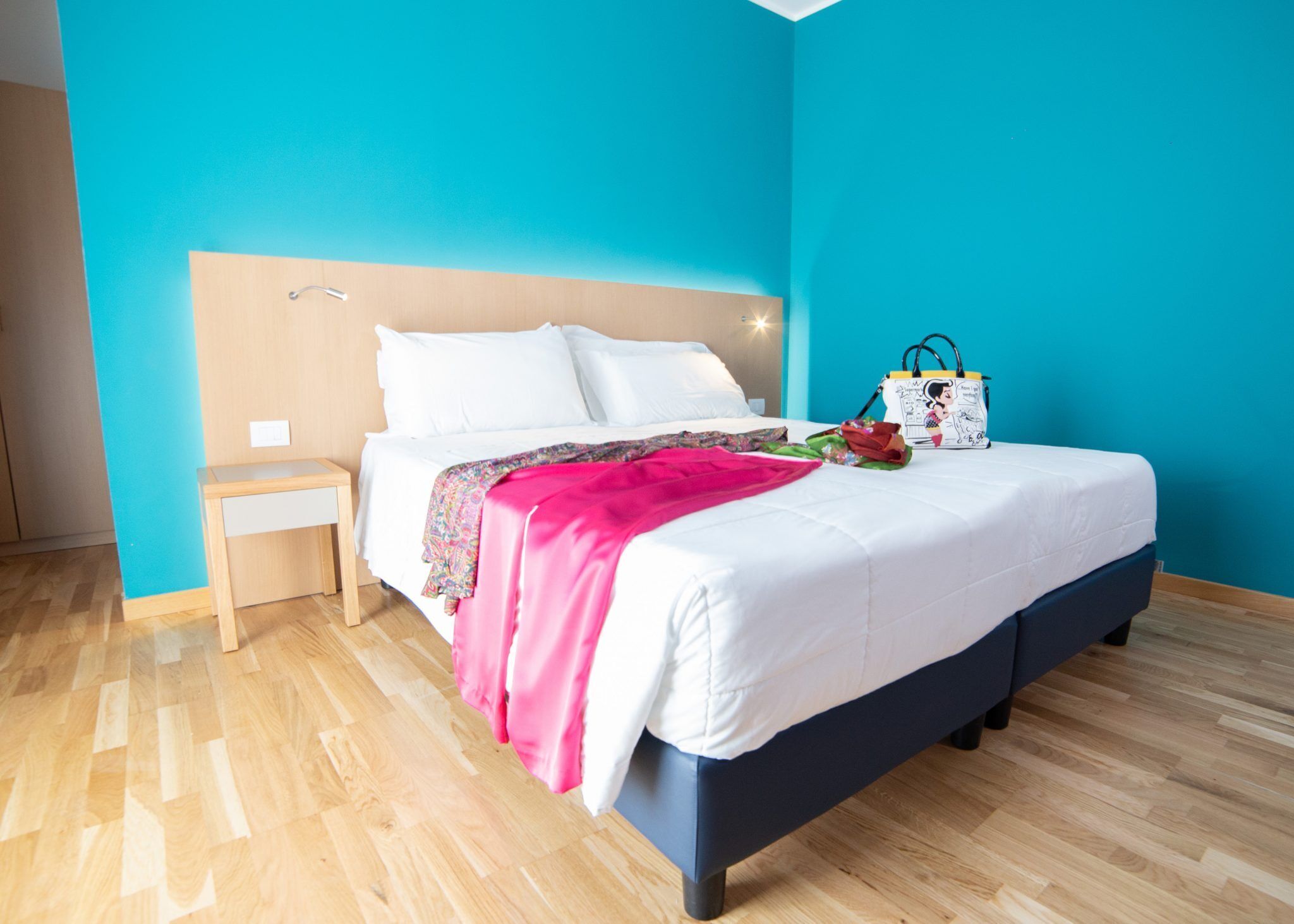 A neatly made bed with white linens and a pink blanket, a bedside table with a lamp, and a wall with a teal color scheme.
