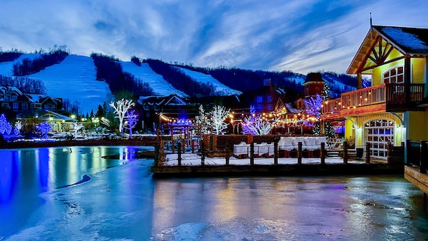 Un hotel de madera con balcón, ubicado junto a un lago con colinas nevadas al fondo.