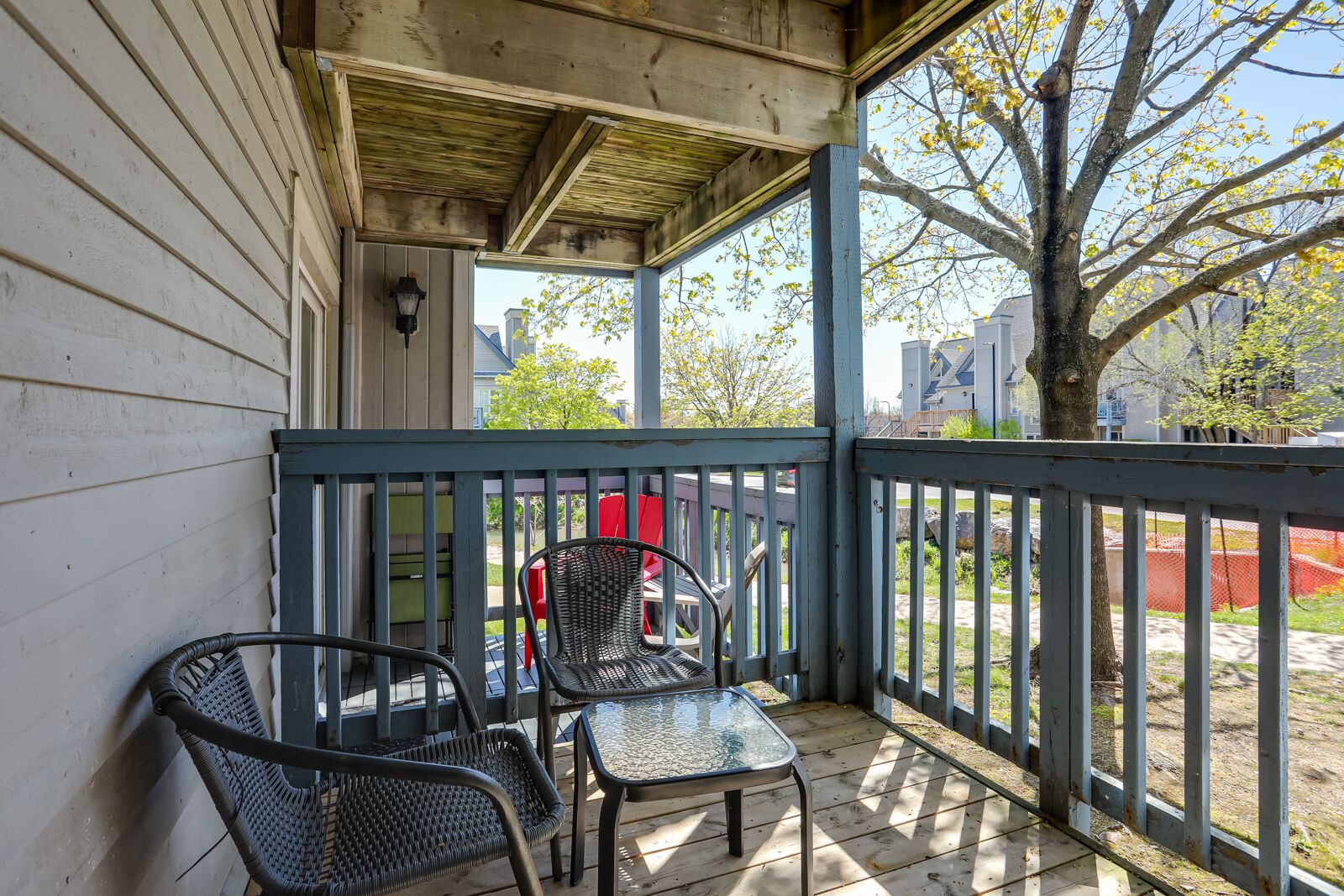 A covered porch with two wicker chairs and a glass table, overlooking a fenced yard with a tree and buildings in the background.