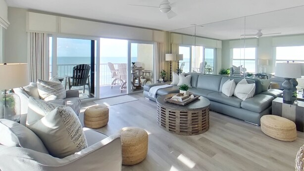 Un salon spacieux avec un canapé gris, des poufs ronds et une table basse. Il y a un balcon avec des chaises et une vue sur l’océan.