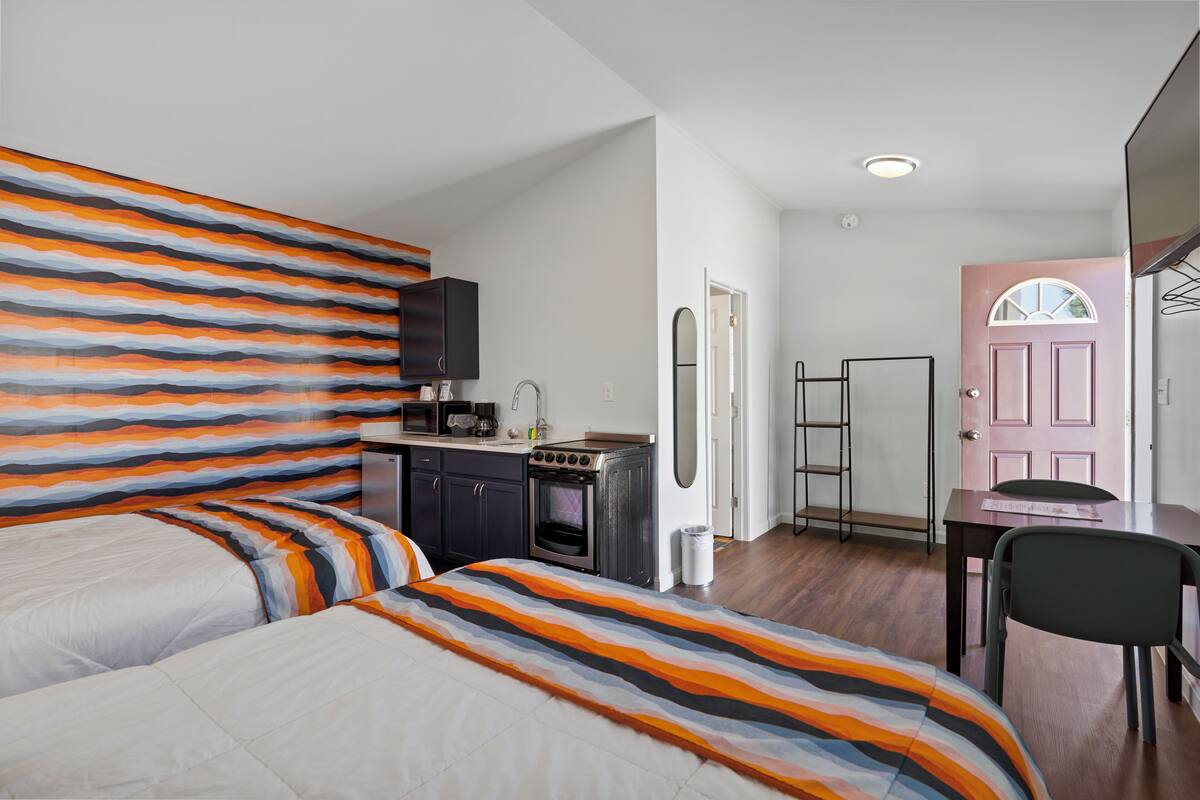 A bedroom with two beds, a kitchenette, a pink door, and a small table with a chair.
