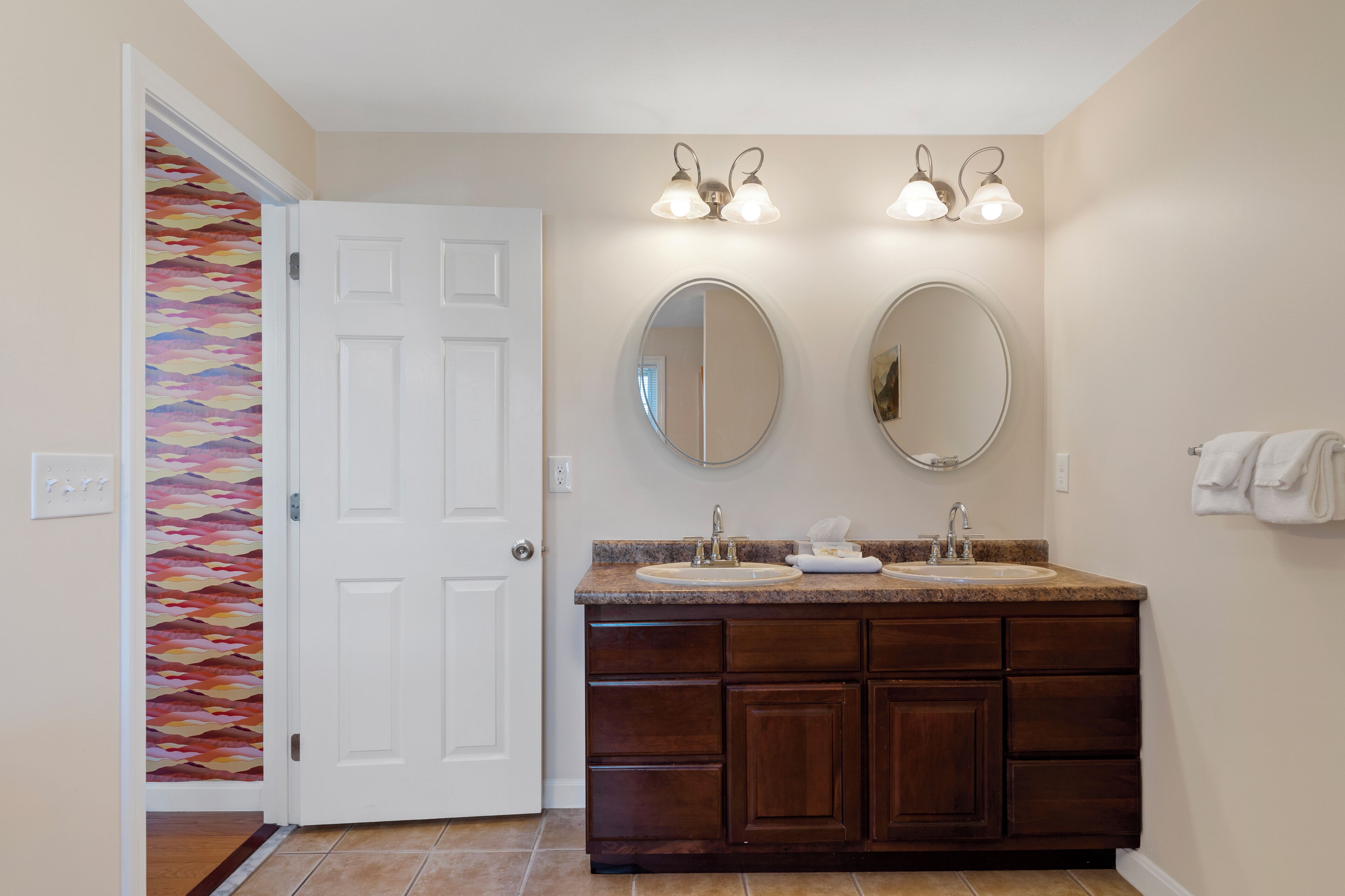 Une salle de bain dotée d’un meuble-lavabo à deux vasques, de deux miroirs fixés au mur et d’une porte donnant sur une autre pièce.