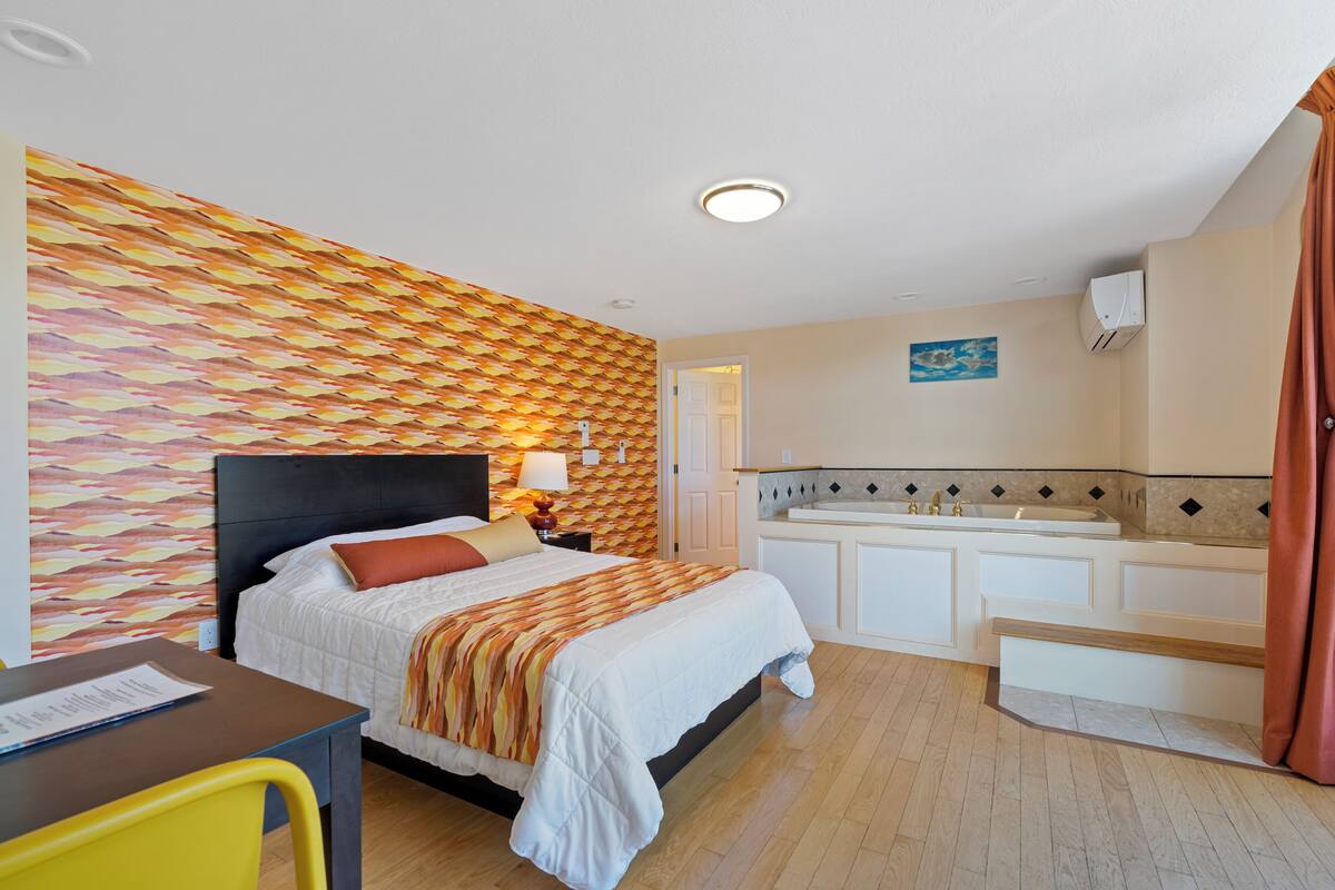 A hotel room with a bed, bedside table, lamp, and a small desk with a chair.