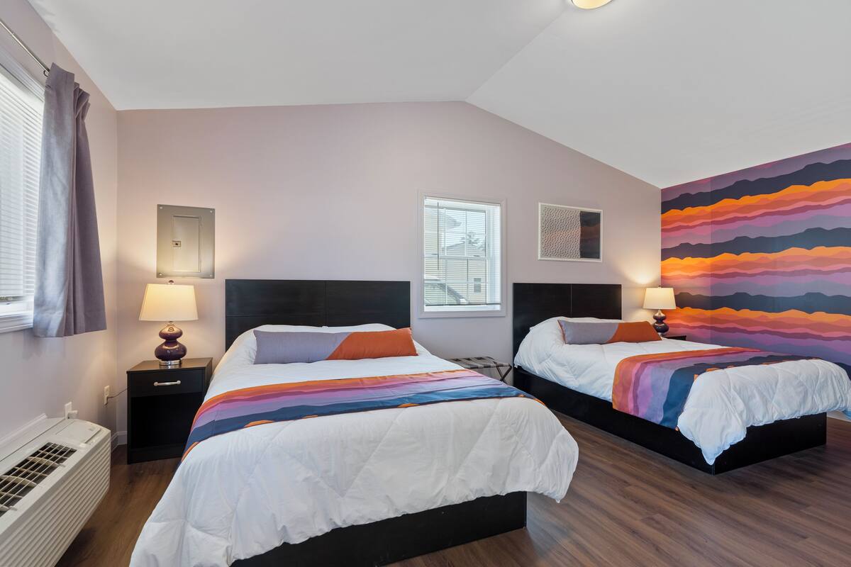 A hotel room with two beds, a large window with blinds, a wall-mounted heater, and a colorful abstract painting.