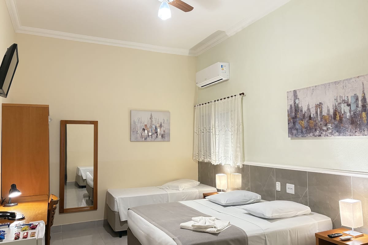 Une chambre d’hôtel avec deux lits, un ventilateur de plafond, un climatiseur, un tableau, un miroir et un bureau avec une lampe et une télécommande.