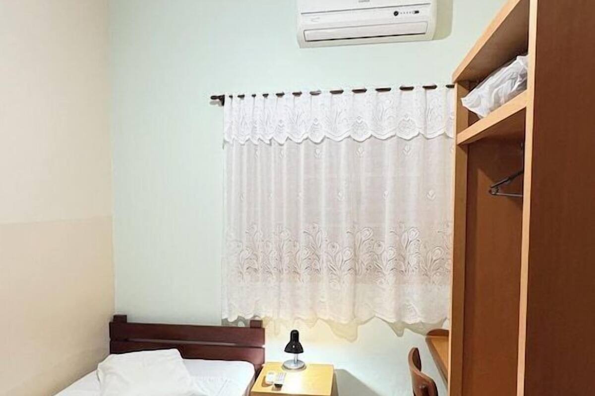 Une chambre avec deux lits simples, un ventilateur de plafond, un appareil de climatisation, une petite table de chevet en bois, une chaise et une armoire.
