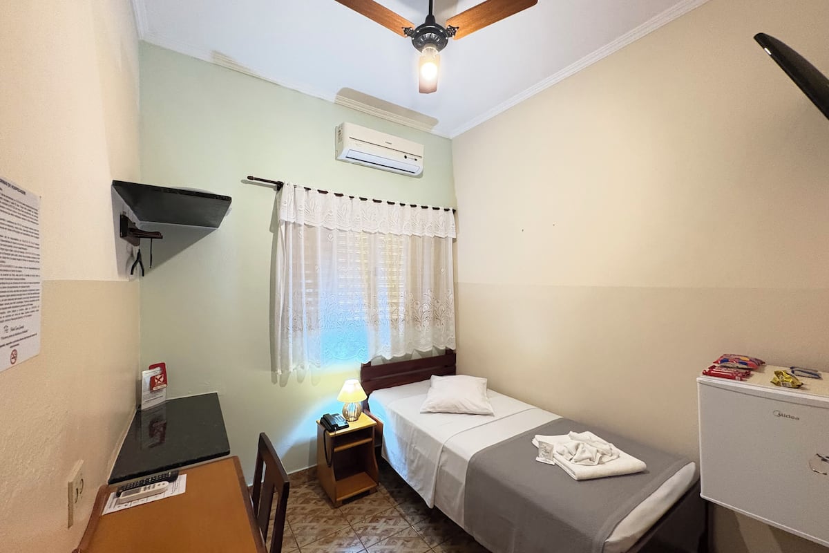 Une petite pièce, bien rangée, comprenant un lit, un bureau, une chaise, un ventilateur de plafond, un climatiseur et un petit placard.