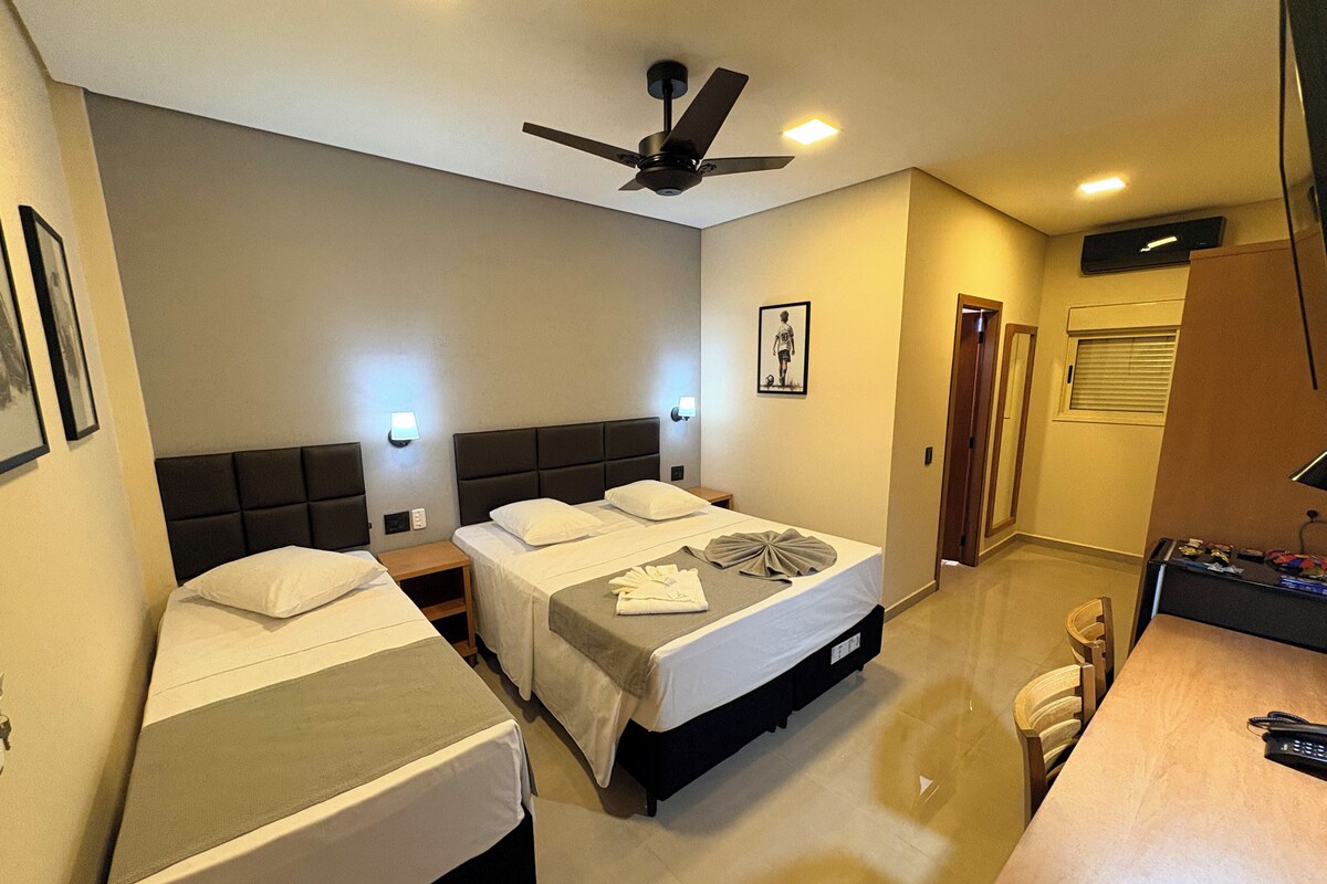 A hotel room with two beds, a desk, a chair, a ceiling fan, and a door leading to another room.