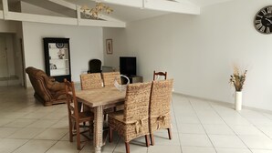 Un comedor con mesa de madera y sillas de mimbre, televisor y un reloj en la pared.