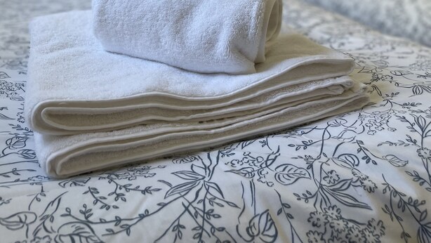 A neatly made bed with a rolled white towel on top, a floral-patterned bedspread, and a bedside table with a lamp.