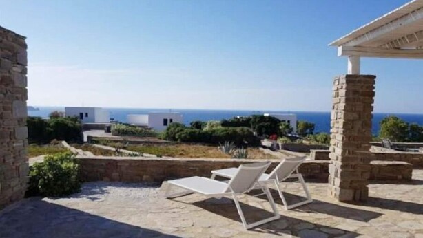 A stone patio with two white lounge chairs and a pergola, overlooking a coastal view with buildings and the sea. - Sunset Villa II (Paros)
