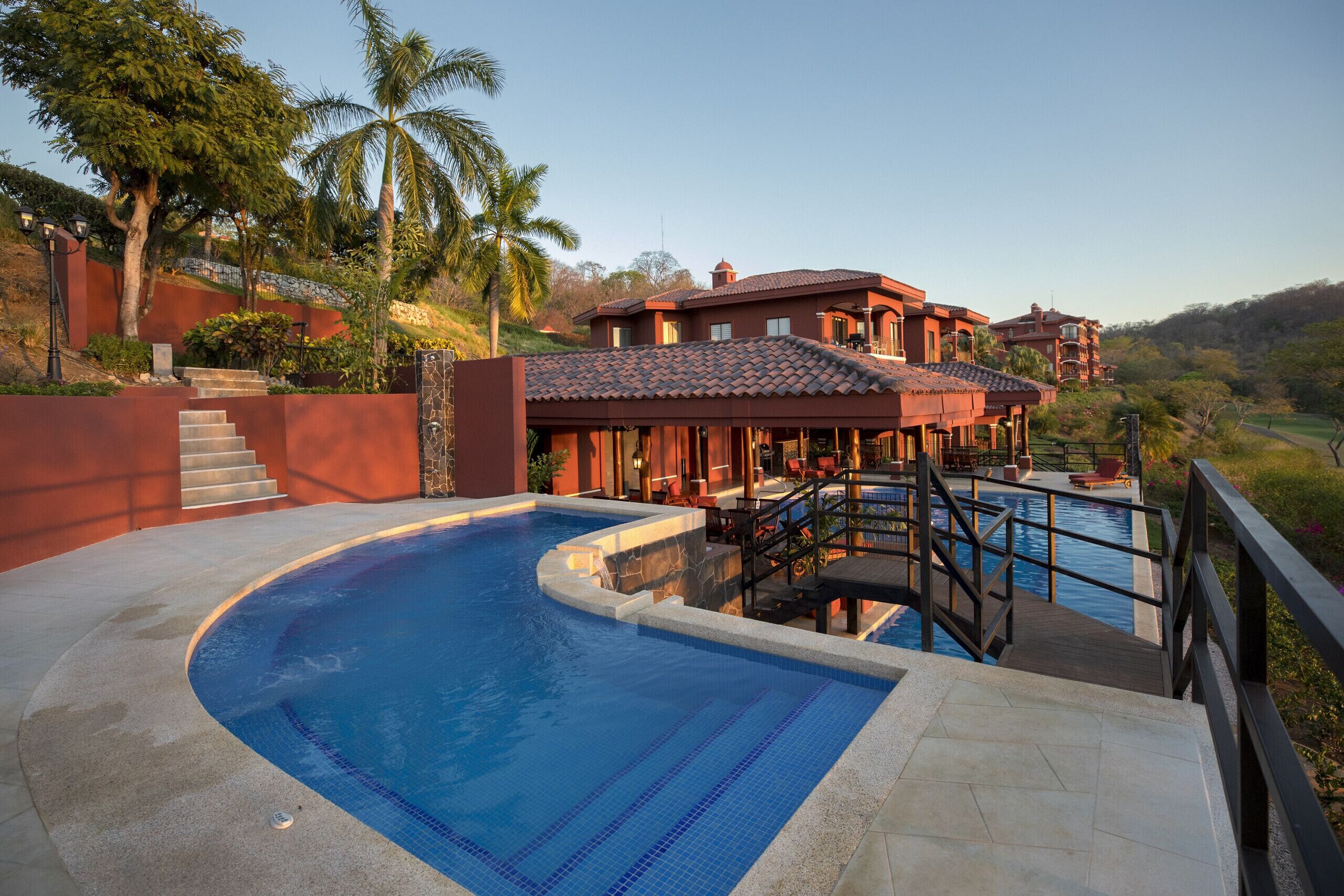 A swimming pool with a curved design, adjacent to a building with a red-tiled roof and a balcony overlooking the pool.