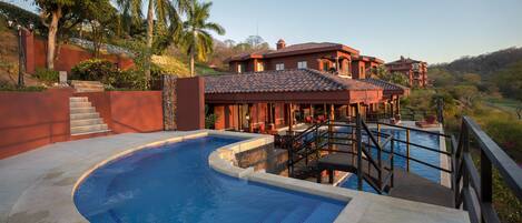 A swimming pool with a curved design, adjacent to a building with a red-tiled roof and a balcony overlooking the pool.