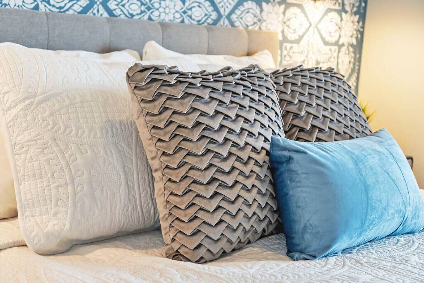 Een bed met een beddengoed met een patroon, een grijs gestoffeerd hoofdbord en decoratieve kussens.