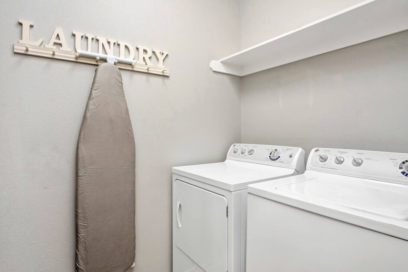 Et vaskerum med to hvide vaskemaskiner, et beige strygebræt og en vægmonteret reol med ordet "LAUNDRY".