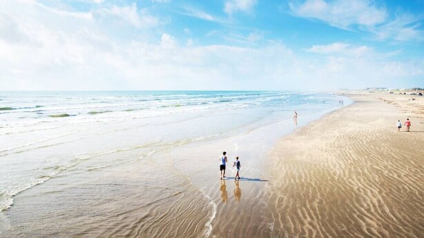 Una playa extensa con olas suaves, cielo despejado y algunas personas caminando por la orilla.
