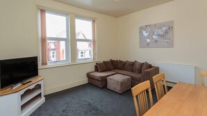 A living room with a large flat-screen TV, a brown sectional sofa, a wooden dining table, and a world map on the wall. - Cherry Property - Coconut Suite (Blackpool)