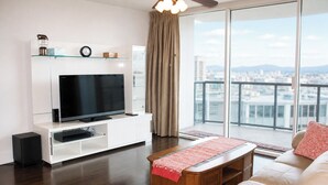A modern living room with a flat-screen TV, white entertainment unit, leather sofa, and a city view through large windows. - Deluxe 2BR Condo in Waikiki! (Honolulu)