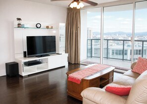 A modern living room with a flat-screen TV, white entertainment unit, leather sofa, and a city view through large windows. - Deluxe 2BR Condo in Waikiki! (Honolulu)
