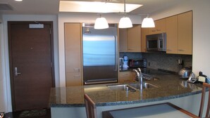 A modern kitchen with a stainless steel refrigerator, a microwave, and a toaster. There is a sink with a faucet, and a wooden door is visible to the left. - Deluxe 2BR Condo in Waikiki! (Honolulu)