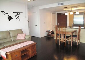 A living room with a sofa, a wooden coffee table, a dining table with chairs, and a kitchen area with a refrigerator and a sink. - Deluxe 2BR Condo in Waikiki! (Honolulu)