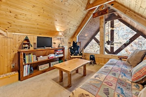 Ein gemütliches Blockhäuschen im Inneren mit Holzbalkendecke, einem Fernseher, Bücherregalen, einem Holztisch, einer Couch und einem großen Fenster mit Blick auf Bäume.