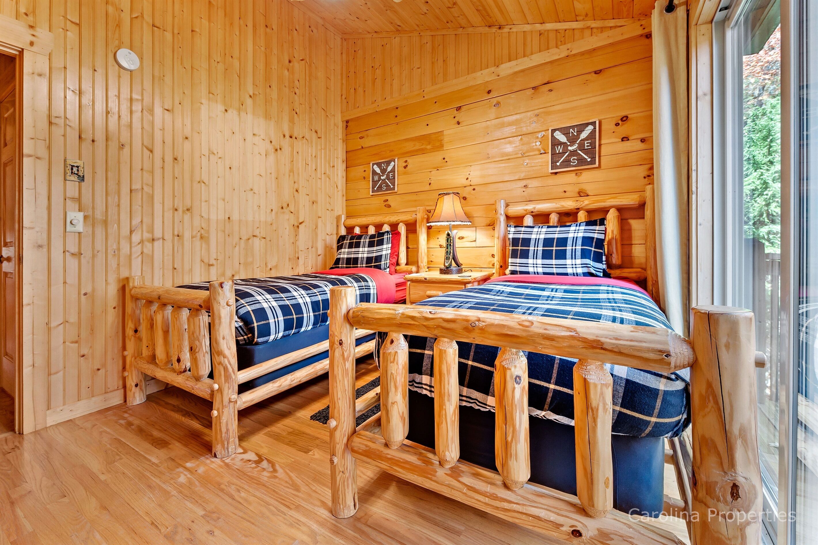 Un dormitorio tipo cabaña de madera con dos camas, una mesita de noche, una lámpara y una ventana con vistas al follaje.