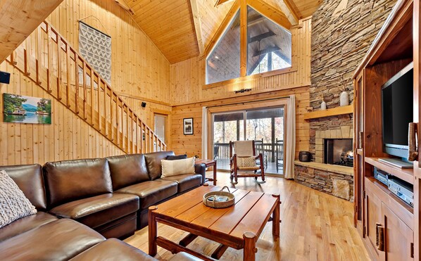 A spacious living room with a wooden staircase, a stone fireplace, and a large window.