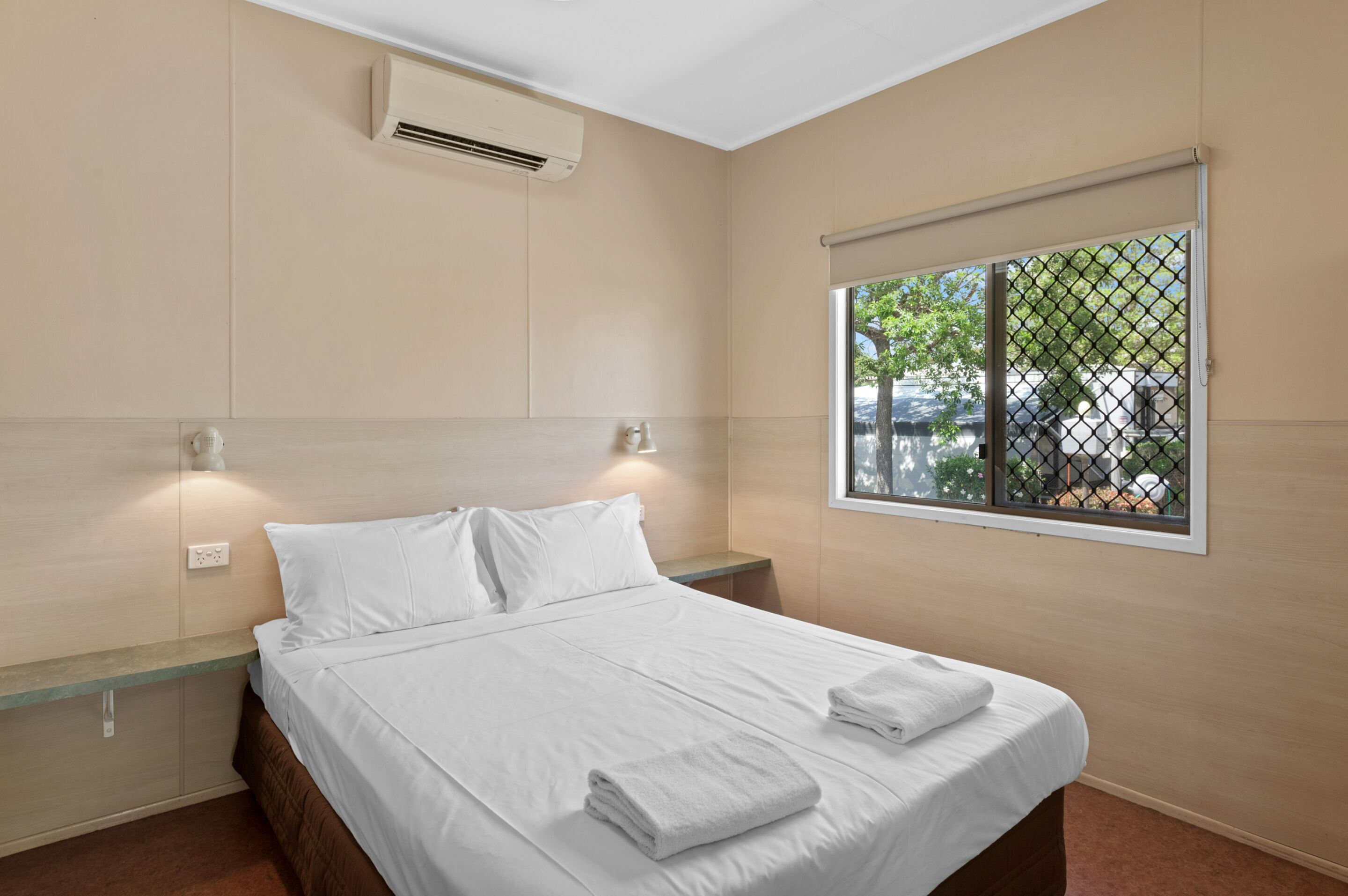 A neatly made bed with white linens and towels, a wall-mounted air conditioner, a window with a view of the outside, and a small bedside table.