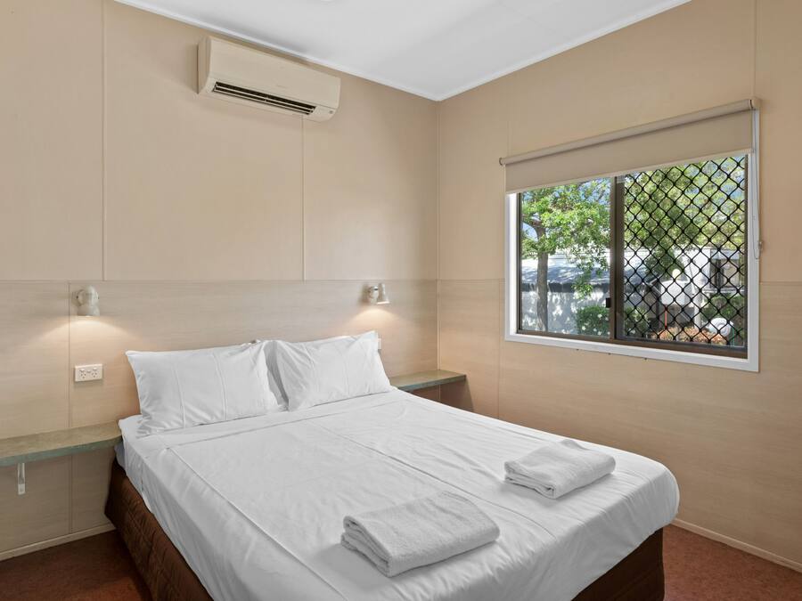 A neatly made bed with white linens and towels, a wall-mounted air conditioner, a window with a view of the outside, and a small bedside table.