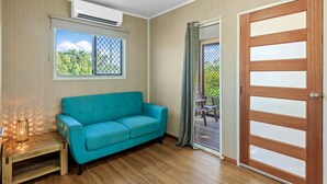 A room with a teal sofa, wooden flooring, a wooden door with glass panels, a window with a view of greenery, and an air conditioning unit. - Discovery Parks - Ayr (Ayr)