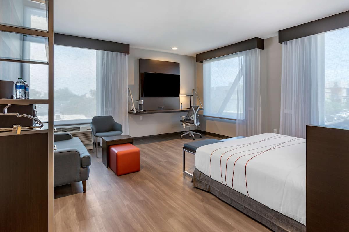 A modern hotel room with a large bed, a desk, a chair, and a sofa. There is a flat-screen TV mounted on the wall, a window with curtains, and a view of the city.