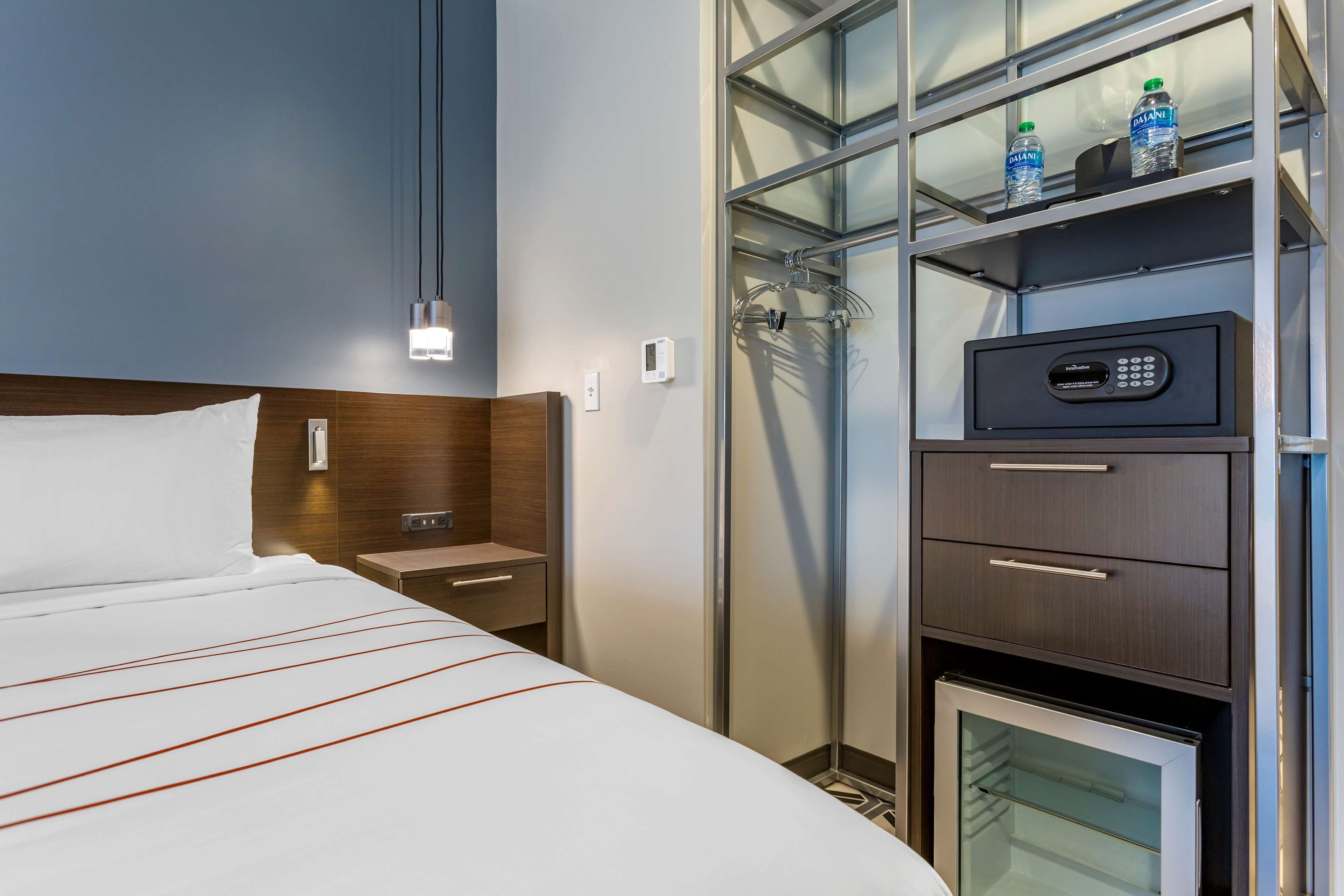 Habitación de hotel con cama, mesita de noche, minibar, zona de ducha con mampara de vidrio y panel de control en un mueble.