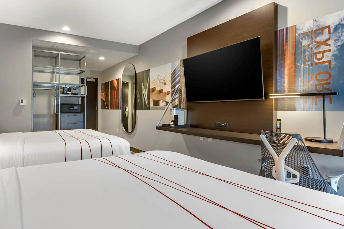 A hotel room with two beds, a TV, a desk with a chair, and a large window.