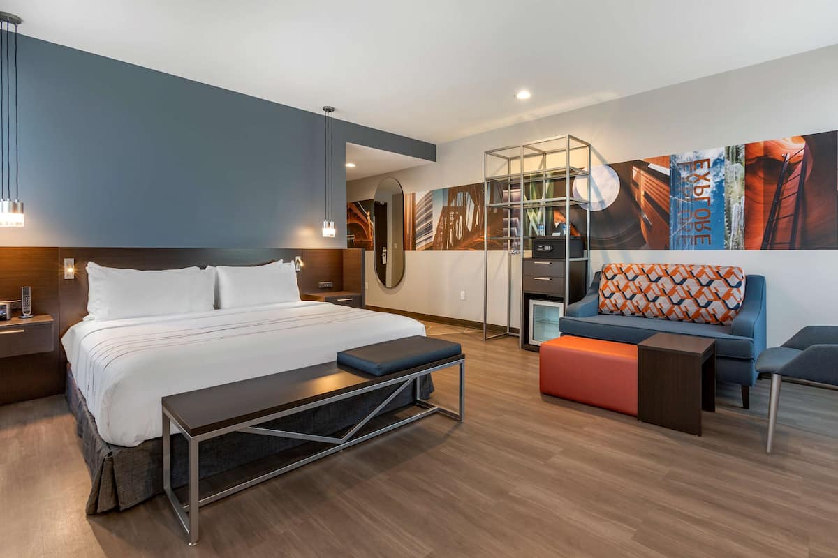 A modern hotel room with a large bed, a sofa, a bench, and a glass partition.