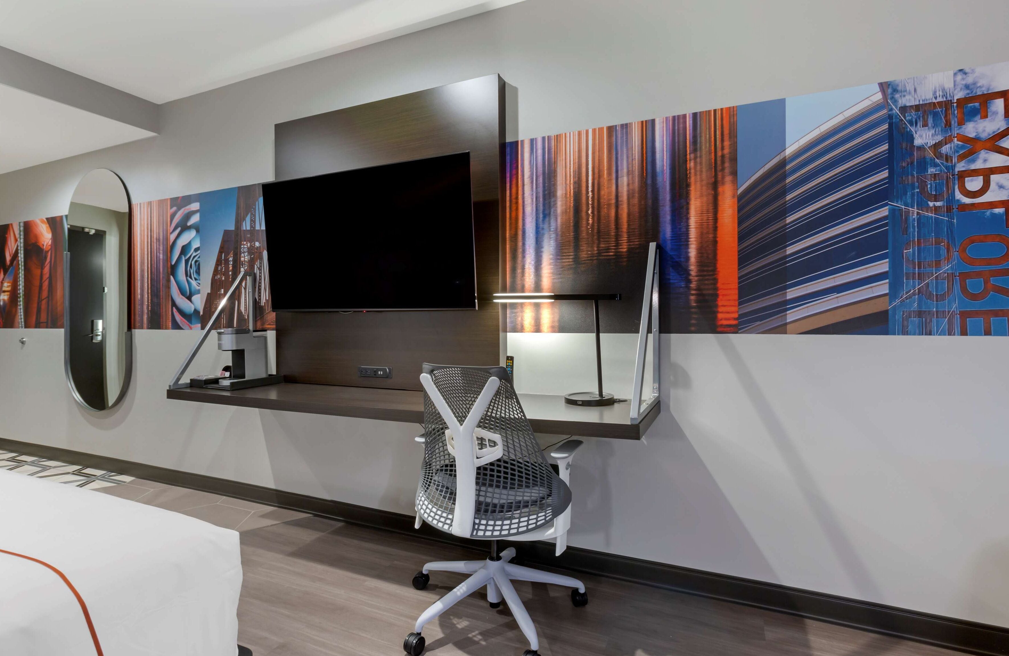 A modern hotel room with a large flat-screen TV mounted on the wall, a desk with a chair, a lamp, and a large mirror.