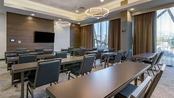 Un salón de conferencias con mesas largas, sillas, un televisor de pantalla plana y ventanas grandes con cortinas.
