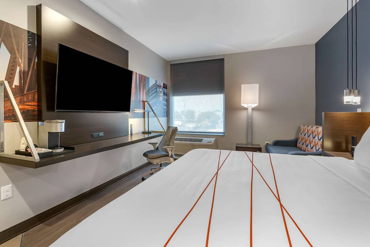 A modern hotel room with a large bed, a flat-screen TV mounted on the wall, a desk with a lamp, and a view of the city through the window.