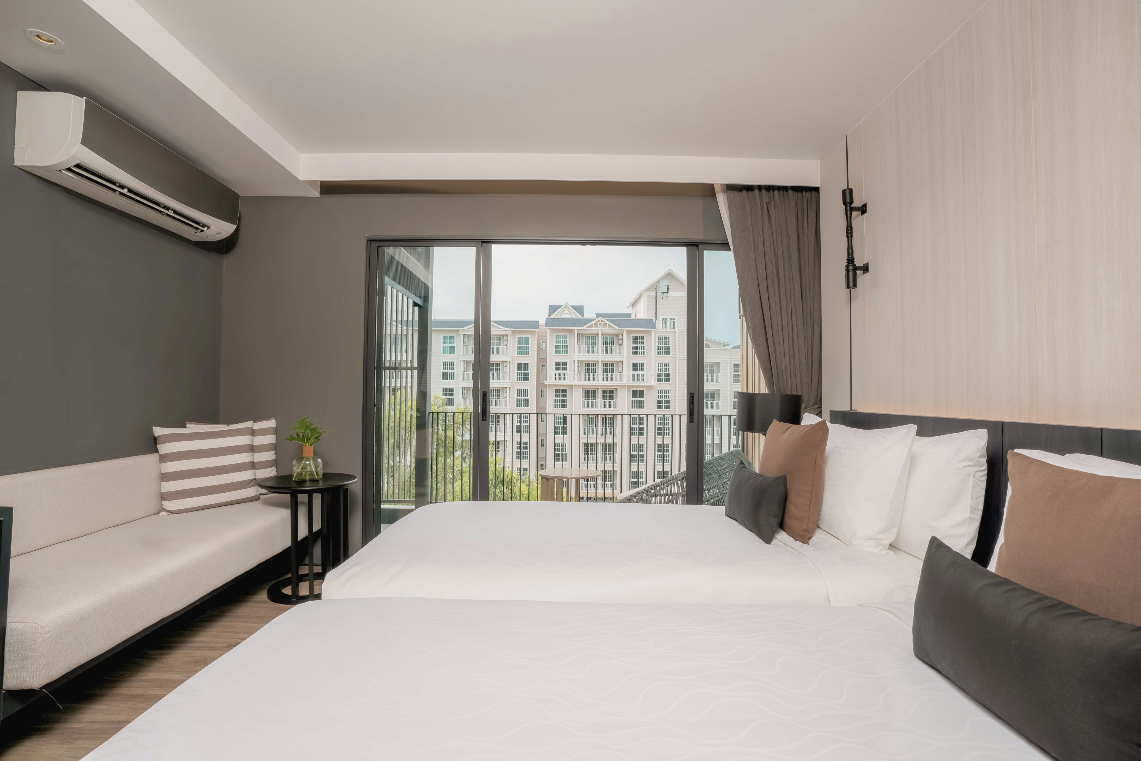 A modern hotel room with a large bed, a sofa, a small table with a plant, and a view of a building through large windows.