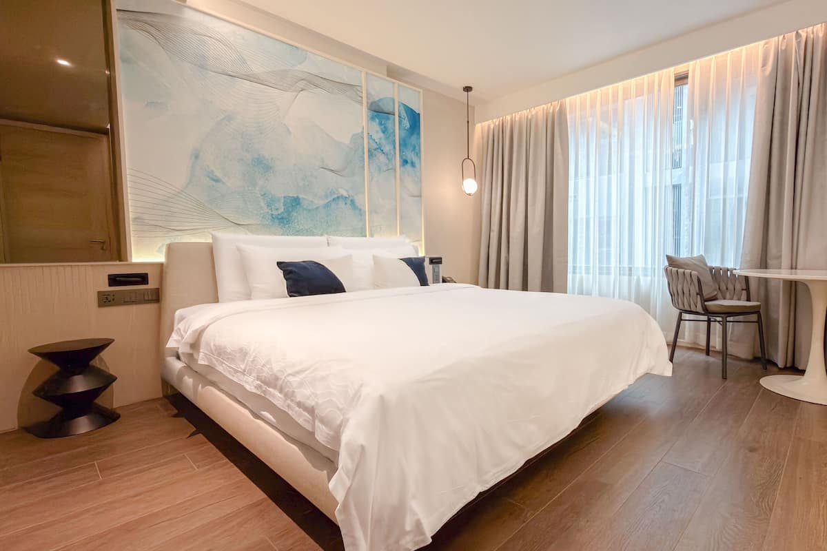 Ein Hotelzimmer mit einem großen Bett, einem Schreibtisch, einem Sessel und einem großen, abstrakten Gemälde an der Wand.