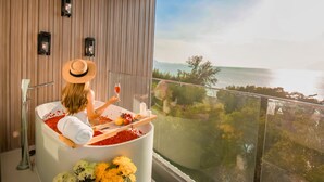 Een vrouw ontspant in een badkuip vol rozenblaadjes, met uitzicht op zee en een balkon met bloemen.