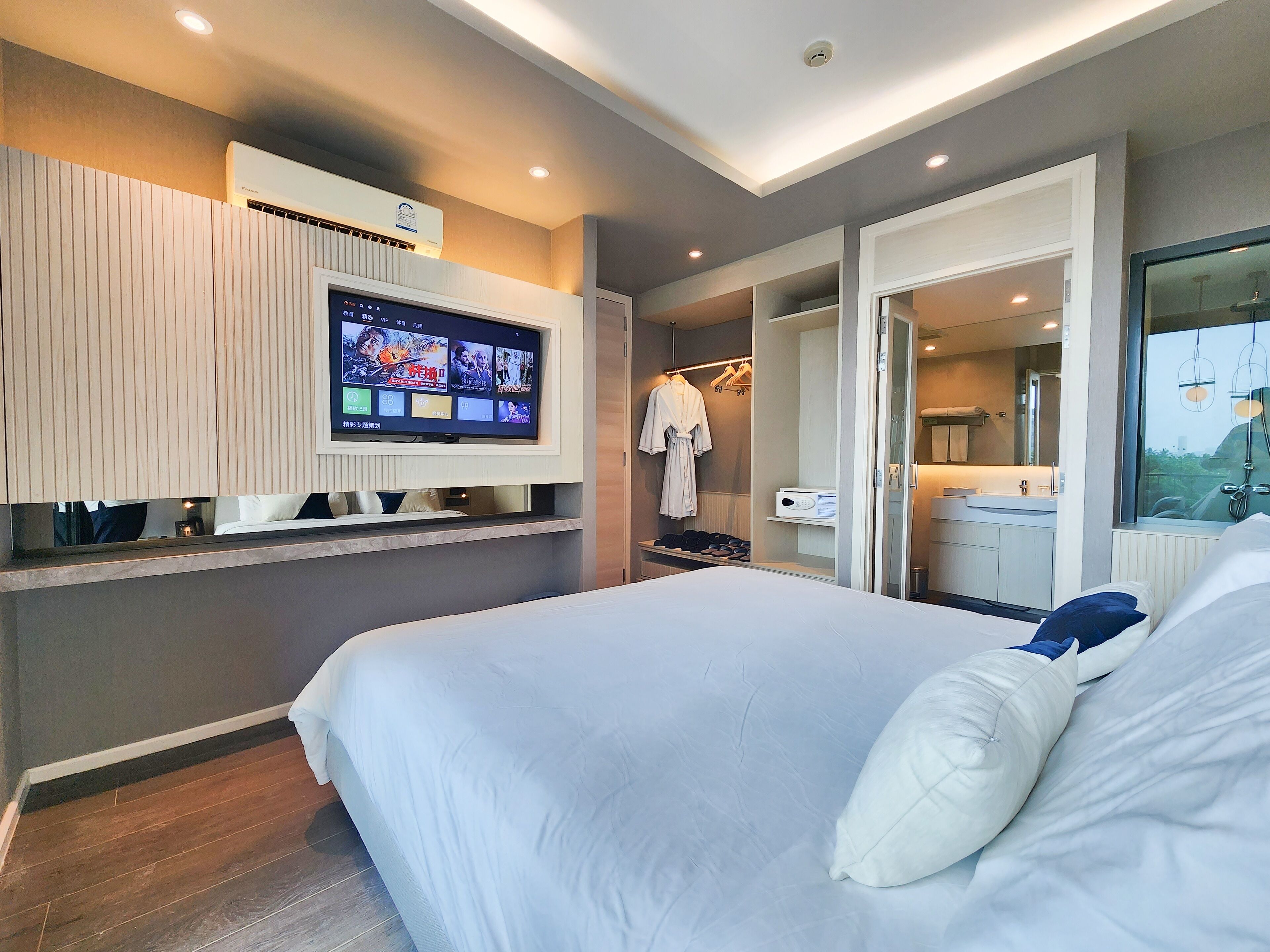 Una habitación de hotel moderna con una cama grande, un televisor montado en la pared, un vestidor amplio y un baño con un espejo grande.